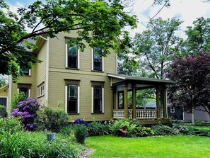 Ein zweistöckiges Haus mit Veranda, umgeben von einem gepflegten Garten mit verschiedenen Pflanzen und Blumen.