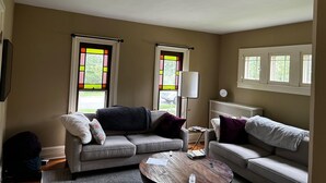 A living room with two sofas, a coffee table, and a lamp. There are windows with colorful stained glass and a clock on the wall.