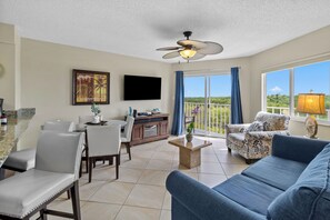 A spacious living area with a ceiling fan, a flat-screen TV, a dining table with chairs, a sofa, and a balcony with a view.