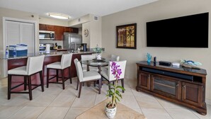A dining area with a round table, chairs, a potted orchid, a TV on the wall, and a kitchen in the background. - 2BR Condo w/ Ocean Views, Private Beach & Pool (Tavernier)