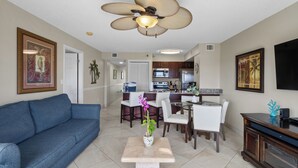 A living room with a blue sofa, a dining table with chairs, a kitchen area, and a ceiling fan. - 2BR Condo w/ Ocean Views, Private Beach & Pool (Tavernier)
