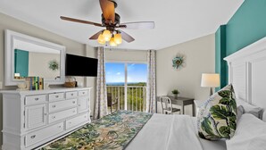 A bedroom with a bed, a desk, a chair, a television, and a balcony with curtains.