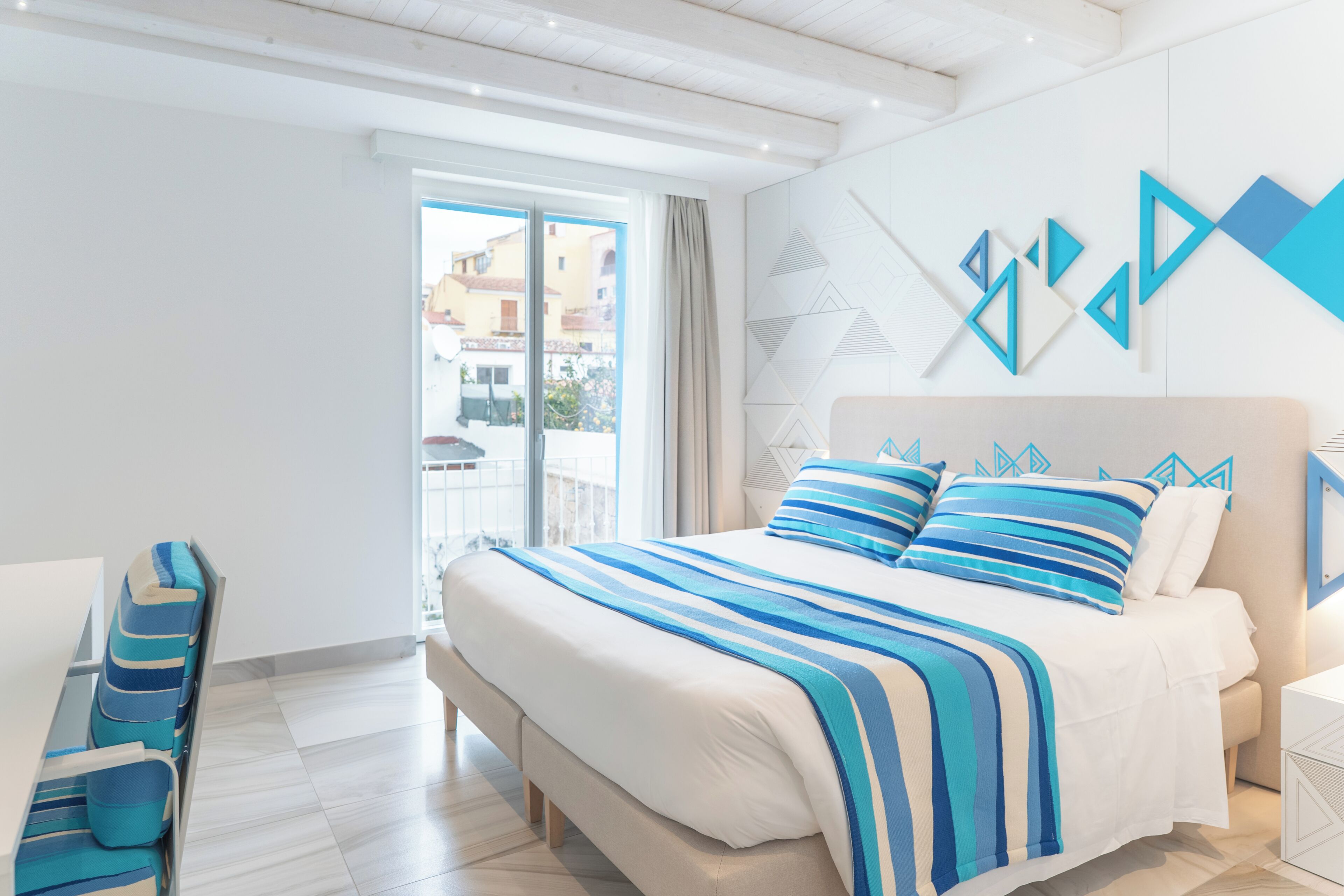 A modern hotel room with a large bed, a seating area with blue-striped chairs, and a view of buildings through the window.