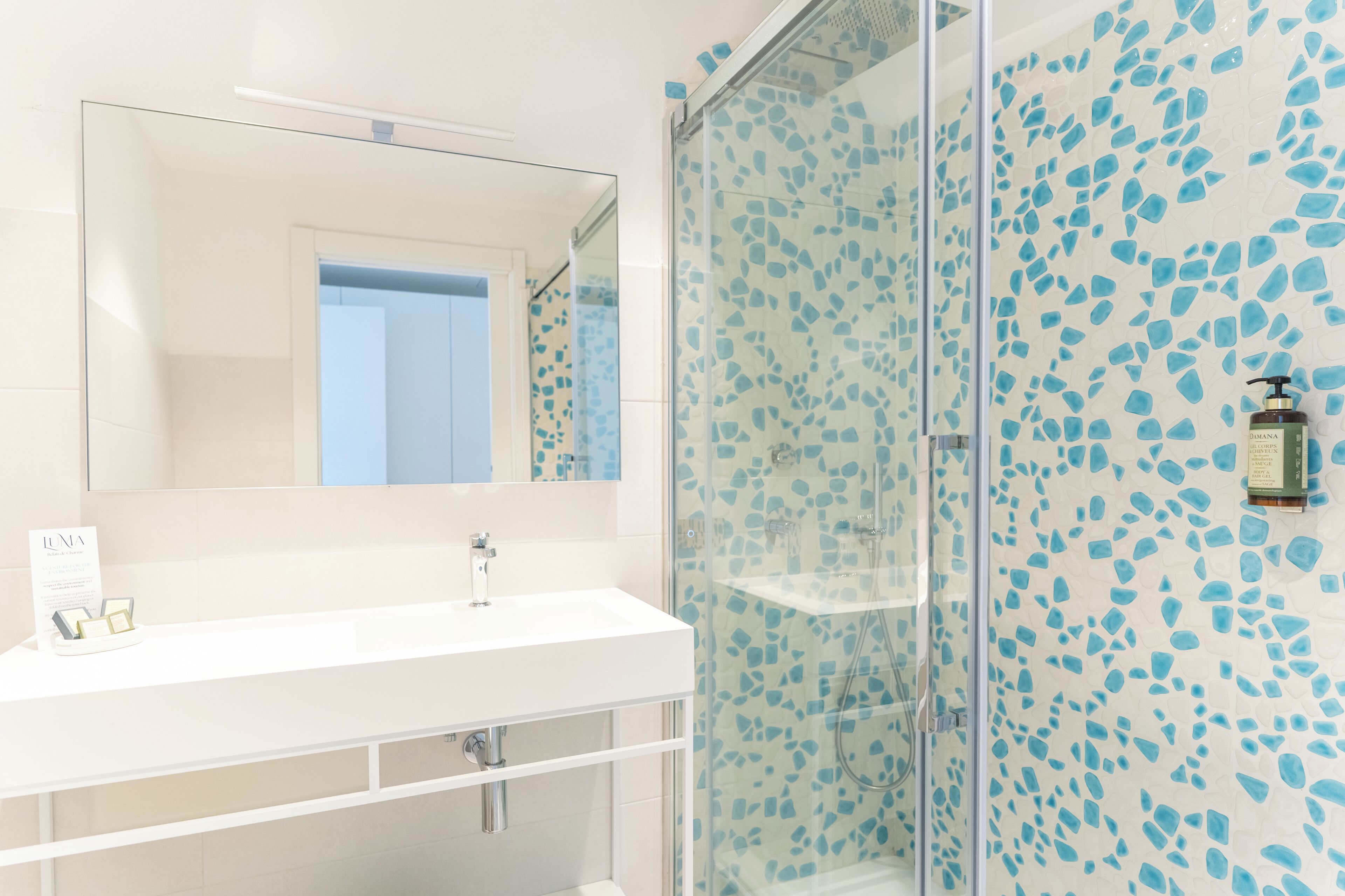Een badkamer met een wit wastafelmeubel, spiegel en een douchecabine met blauwe mozaïektegels.