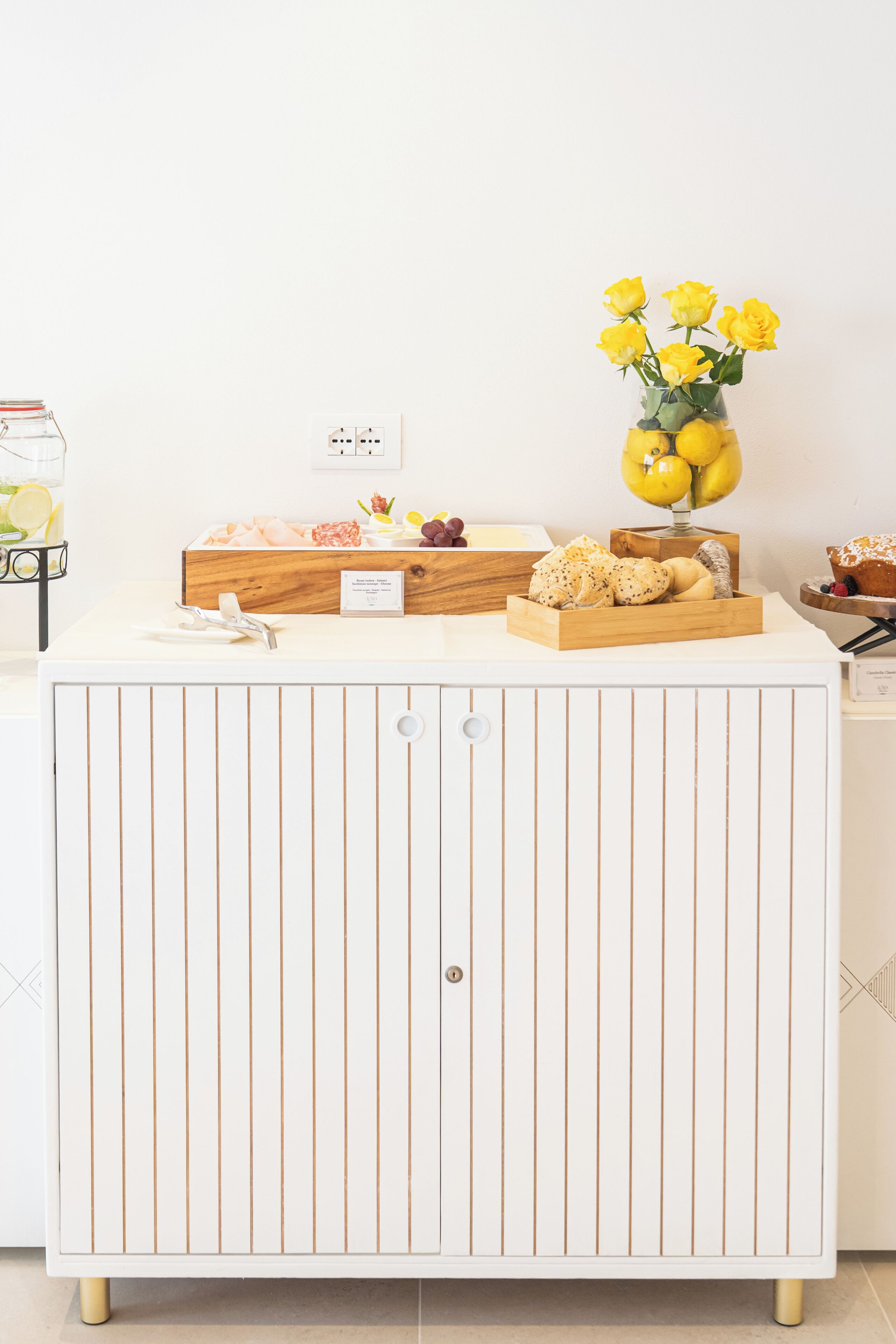 A white cabinet with vertical lines, a wooden tray with bread, a vase with yellow flowers, and a small sign.