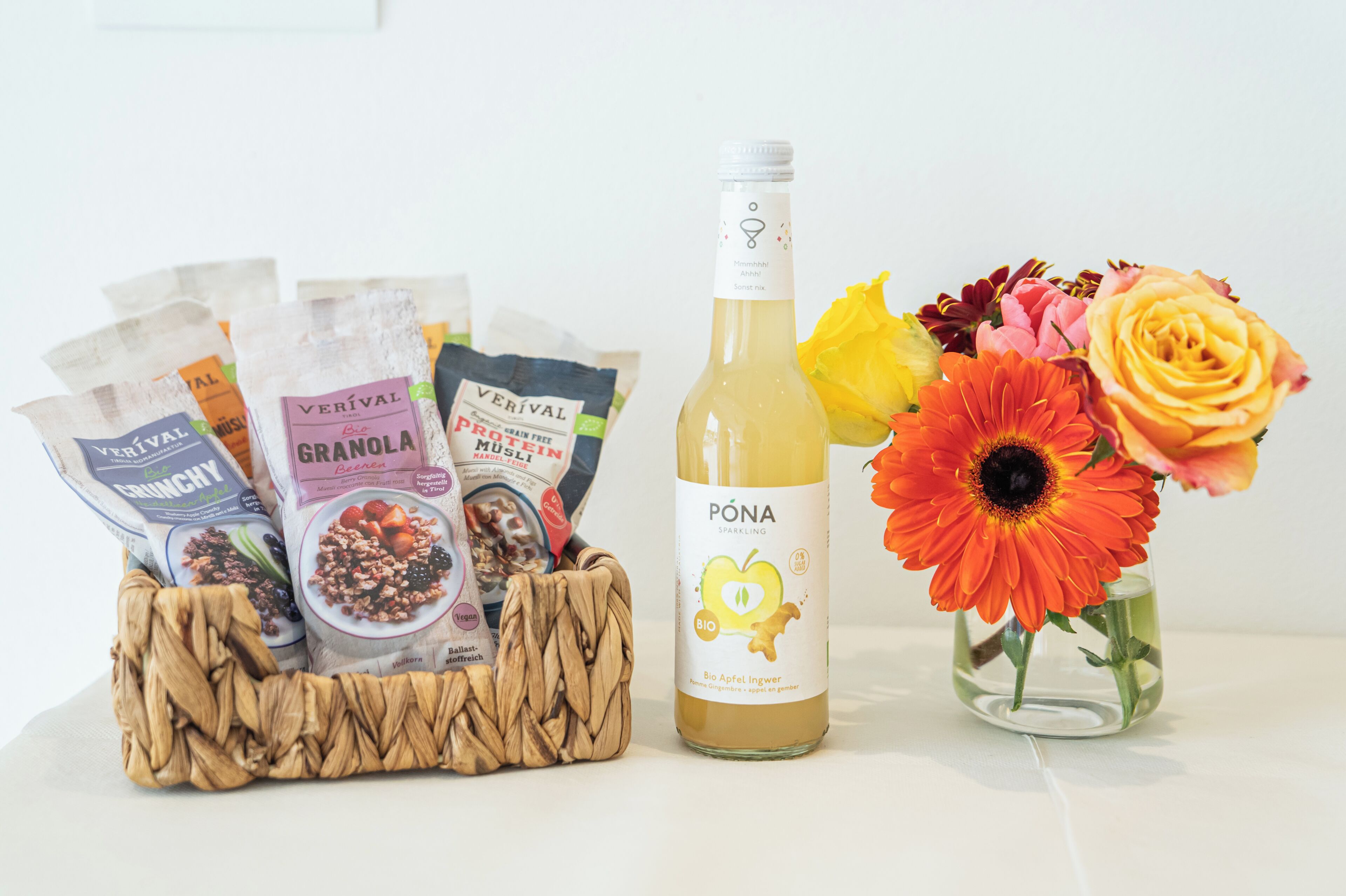 A bottle of Póna sparkling wine, a basket of granola bars, and a vase with flowers.