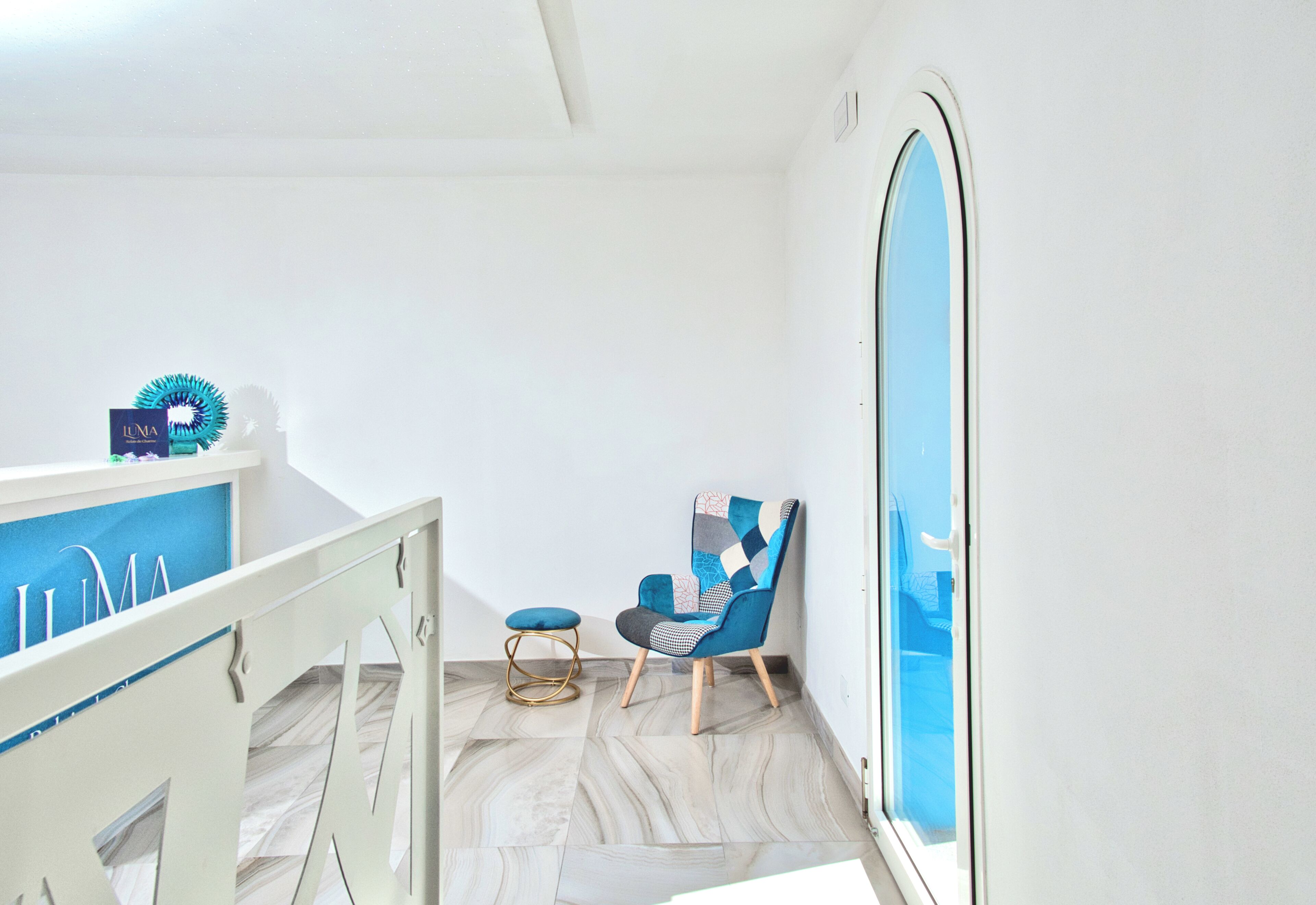 Una habitación con pared blanca, un ventanal grande, una silla azul, un taburete pequeño y un armario blanco con detalles azules.
