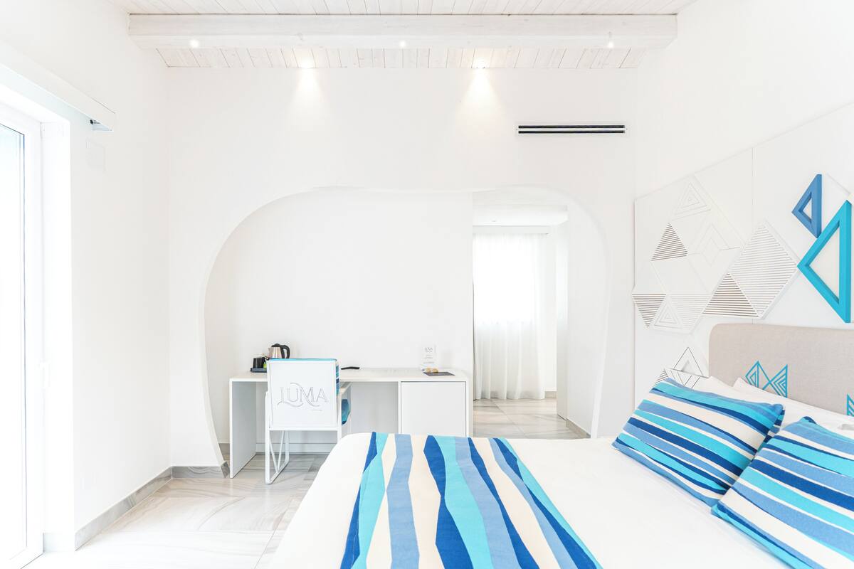A modern bedroom with a bed, a desk, and a chair. There is a window and a door.