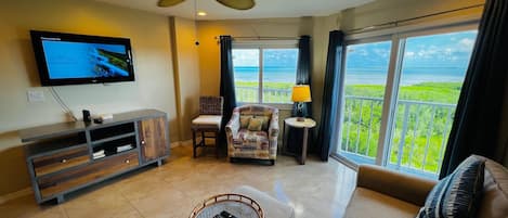 A spacious living room with a large TV, comfortable seating, and a balcony with a view of the ocean.