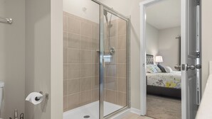 A modern bathroom with a glass-enclosed shower, a toilet, and a visible bed in the adjacent room.