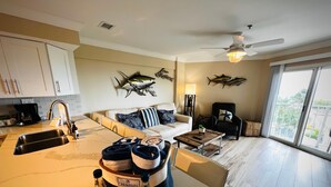 En moderne stue med sofa, sofabord og spiseplads med vask og komfur. Dekorative fisk pryder væggen, og der er udsigt til grøn bevoksning udenfor.