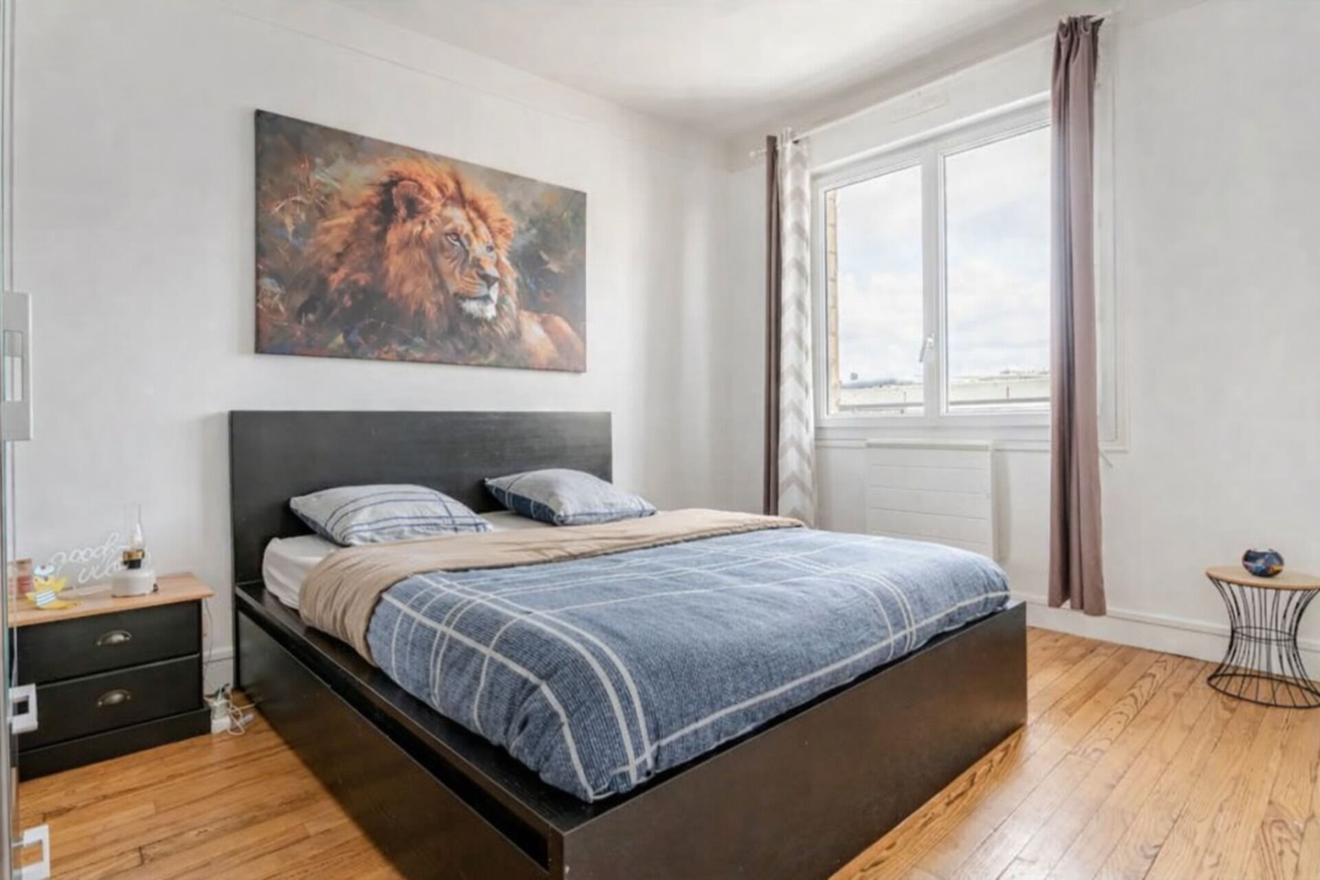 Ein Schlafzimmer mit einem Bett, einem Nachttisch, einem Fenster mit Vorhängen und einem Bild an der Wand.
