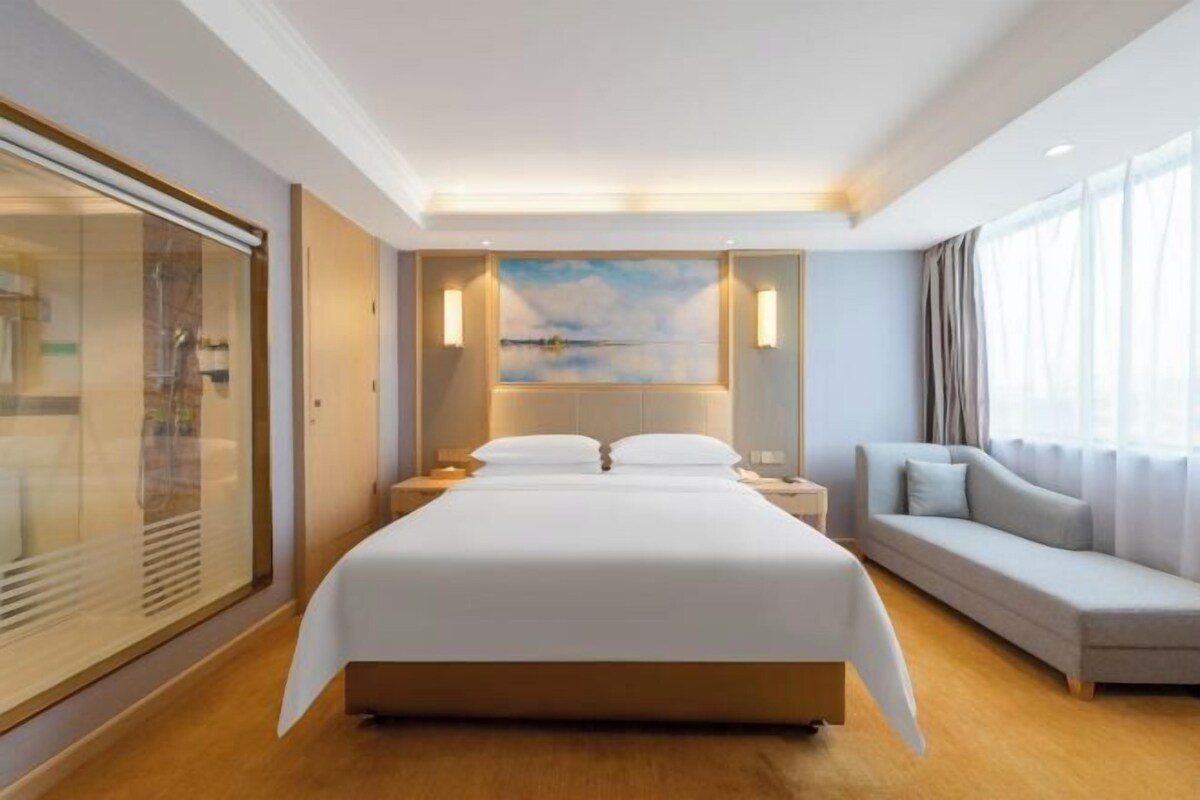 A hotel room with a large bed, a sofa, a painting on the wall, and a view of the outdoors.