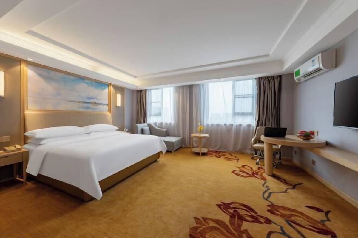 Ein Hotelzimmer mit einem großen Bett, einem Schreibtisch mit Stuhl, einem kleinen Tisch mit Lampe, einem Fernseher und einem Fenster mit Vorhängen.