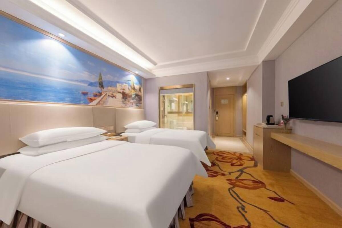 Ein Hotelzimmer mit zwei Betten, einem großen Gemälde mit Strandmotiv, einem Fernseher und einem Teppich mit floralem Muster.