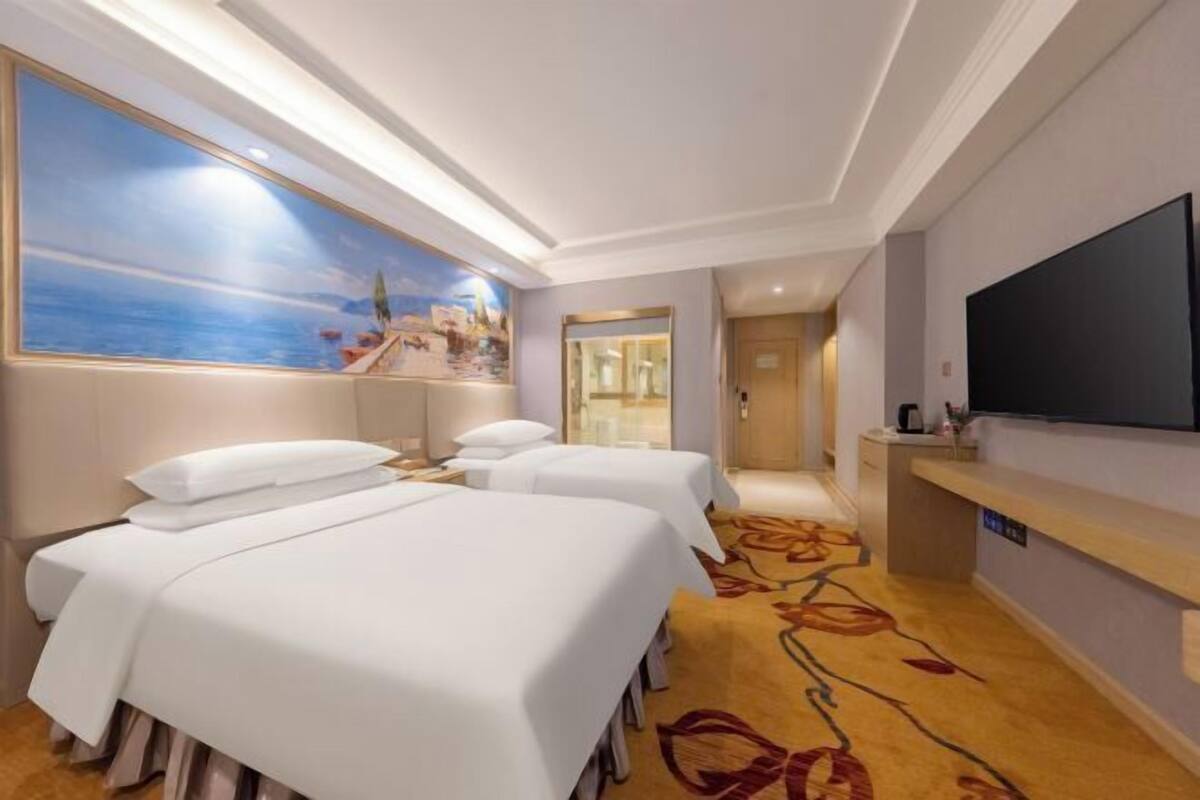 Ein Hotelzimmer mit drei Betten, einem großen Gemälde mit Strandmotiv, einem Flachbildfernseher und einem Empfangstresen.
