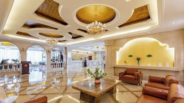 A spacious lobby with a marble floor, leather sofas, a central coffee table with a plant, and a reception area with a yellow wall and a chandelier. - Vienna Hotel (Renhe subway station store of Guangzhou Baiyun International Airport) (Guangzhou)