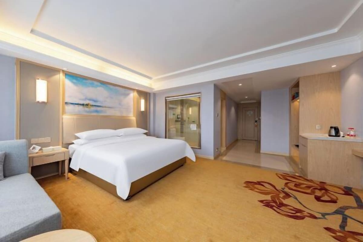 A hotel room with a bed, a desk, a chair, a nightstand, a mirror, and a bathroom visible through an open door.