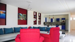 Una sala moderna con un sofá rojo, un sofá gris y una mesa de centro blanca. Hay una cocina con armarios azules y una encimera blanca.