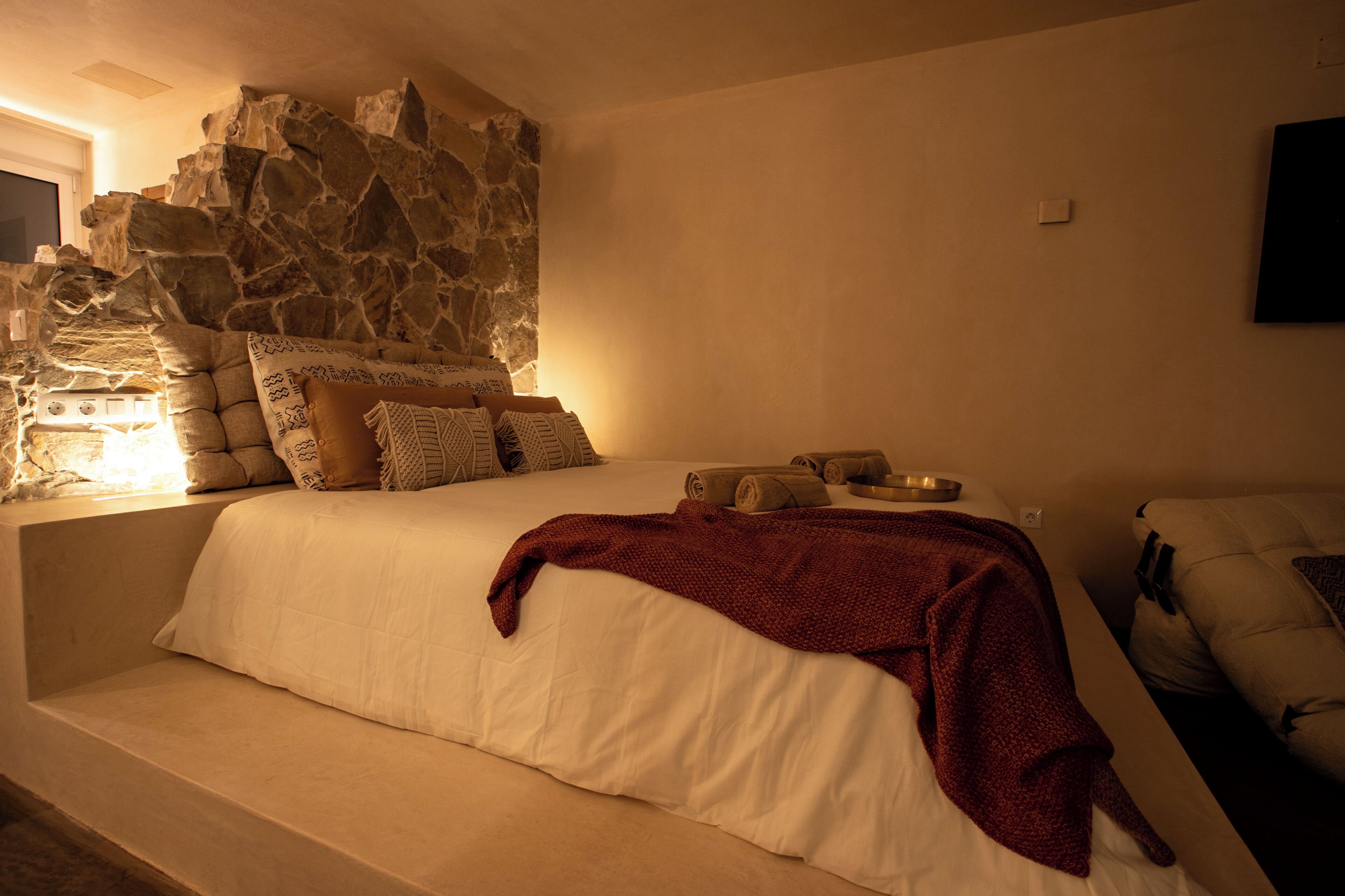 A bedroom with a stone wall, a bed with white bedding, and a red blanket.