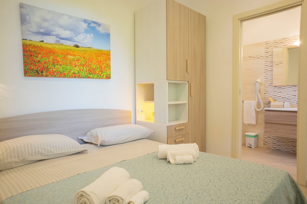 Habitación de hotel con cama, almohadas, un cuadro, armario, espejo y baño visible a través de una puerta abierta.