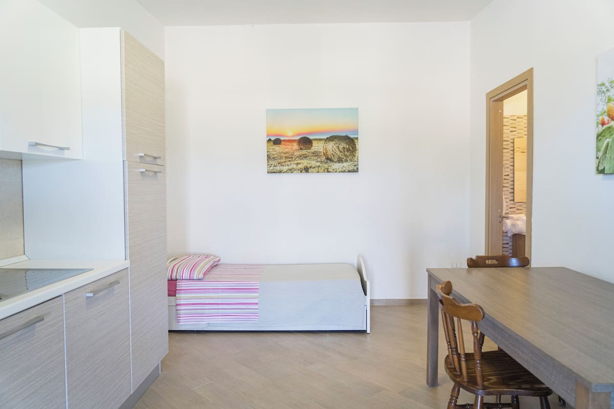 Habitación pequeña con cama, mesa de comedor y una silla. Hay un cuadro en la pared y una puerta que da a otra habitación.