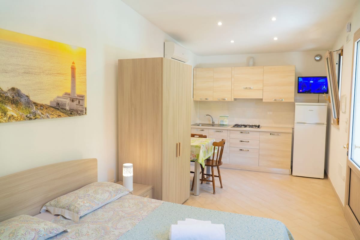 Un dormitorio con cama, un mesita de noche, una lámpara en la mesilla, un cuadro de un faro y vistas a una cocina con mesa y sillas.