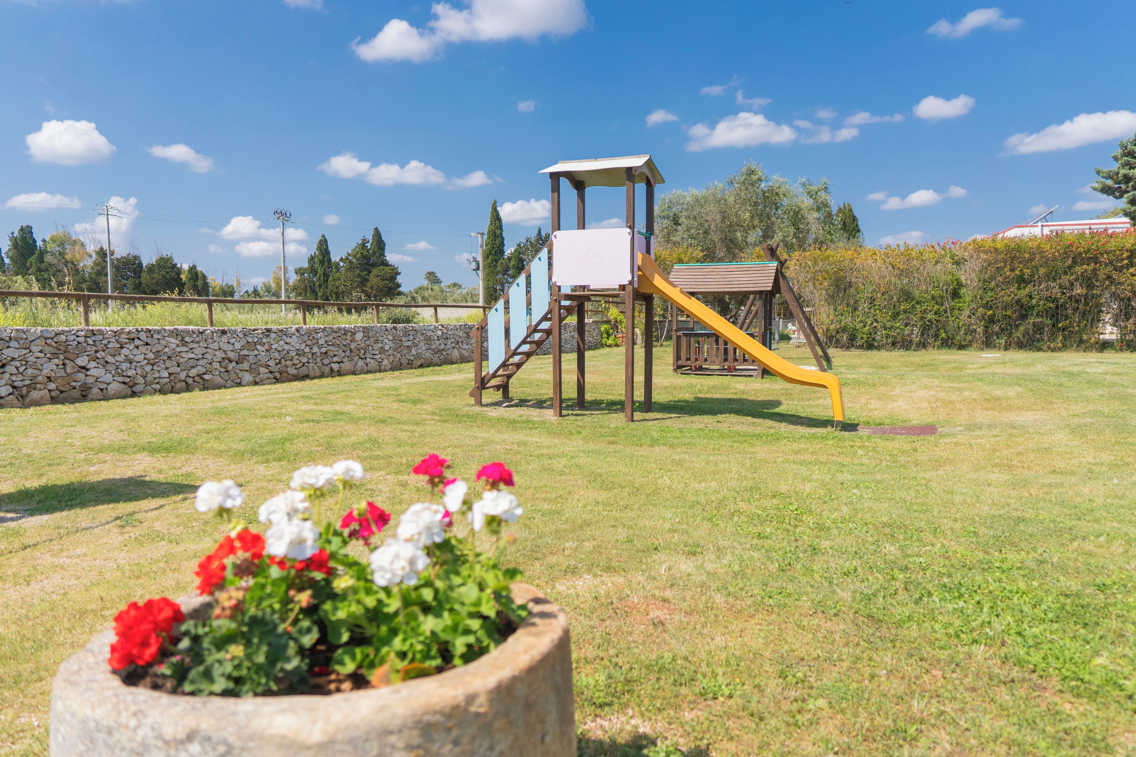 Un parque infantil con tobogán y estructura para escalar, rodeado de un muro de piedra y vegetación.