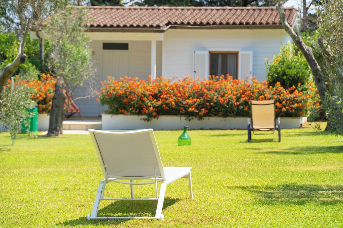 Una silla blanca en un área con césped, con una casa al fondo y un parterre de flores anaranjadas.