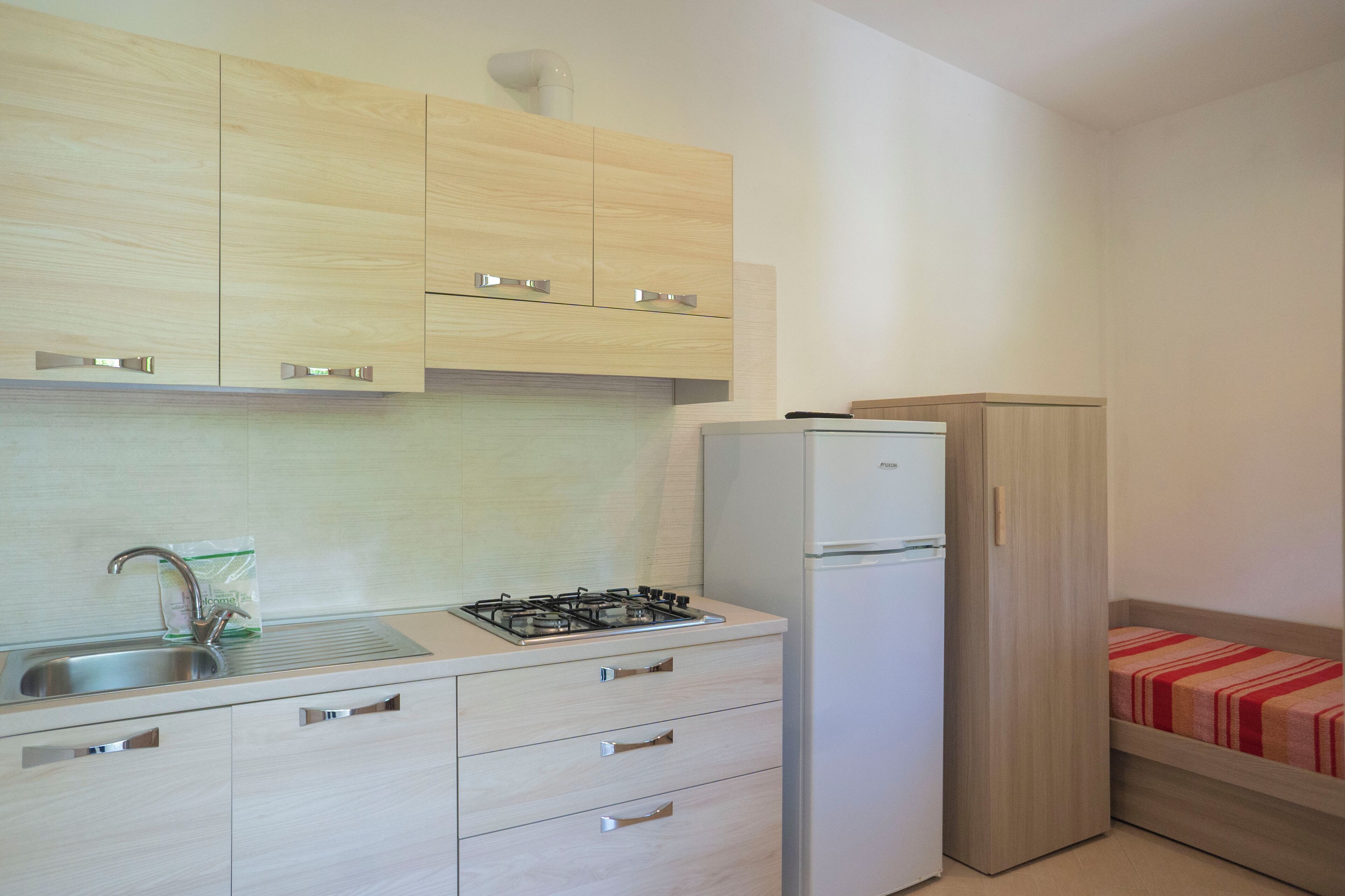 A compact kitchen with a gas stove, sink, and a refrigerator. There is a wooden cabinet above the stove and a small bed with a red blanket in the corner.