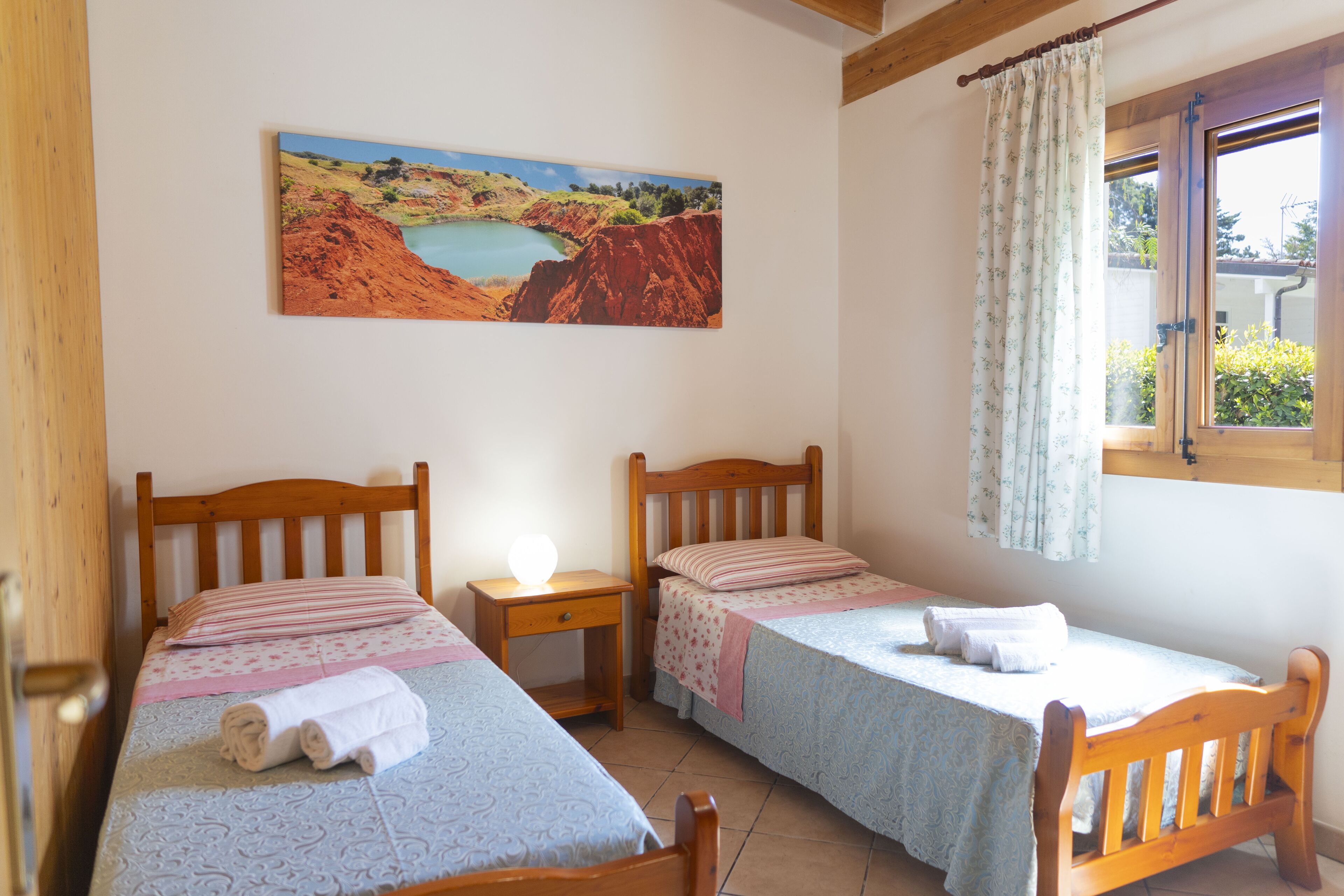 Ein Zimmer mit zwei Holzbetten, einem Nachttisch, einem Landschaftsbild und einem Fenster mit Vorhängen.