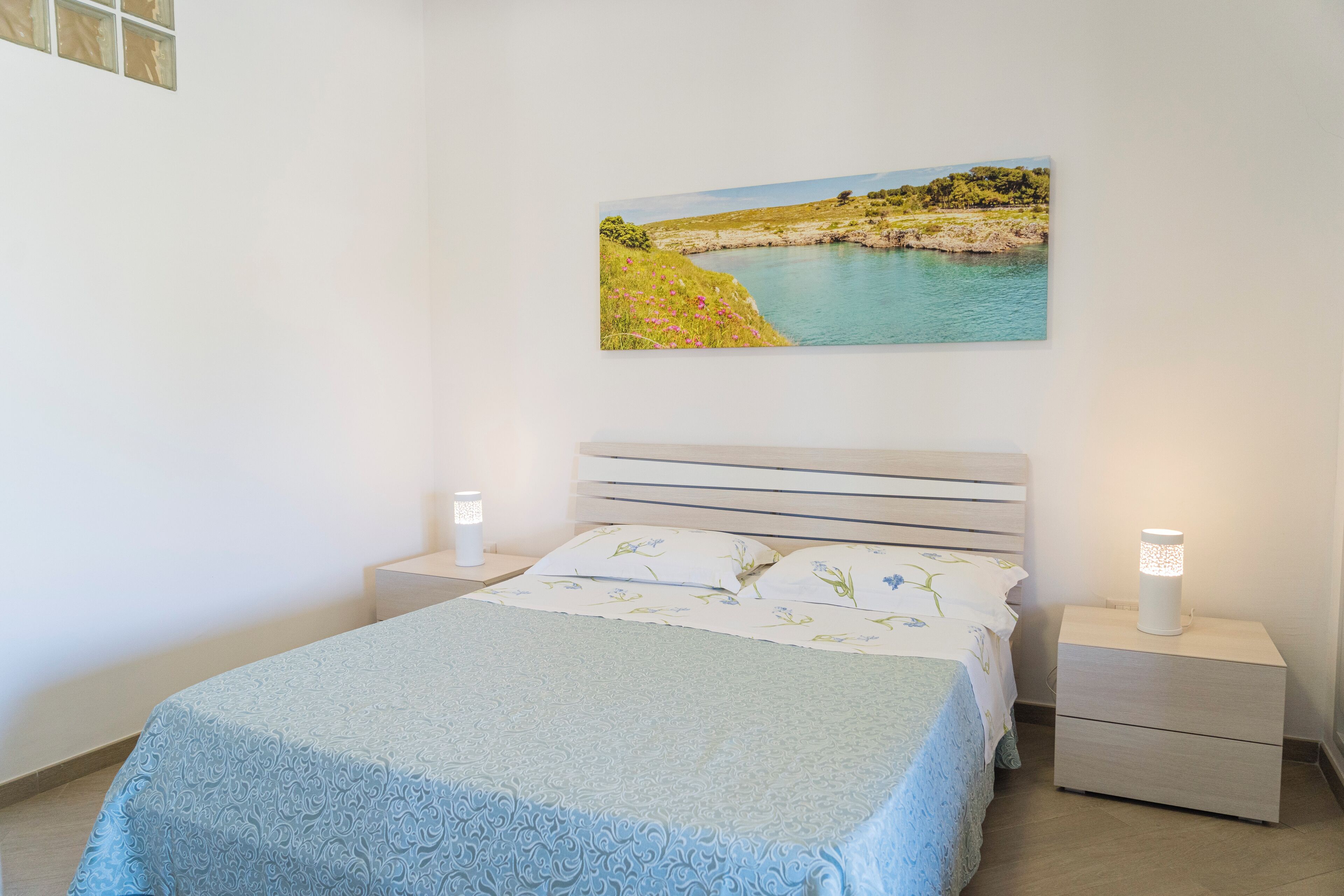 A bedroom with a bed, a nightstand, a bedside lamp, and a wall-mounted picture of a landscape.