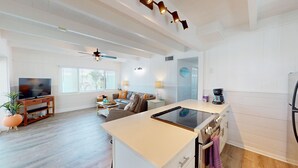A spacious living area with a TV, sofa, and dining table. A kitchen with a stove and coffee maker is visible. - Oceanview 3-bdrm home steps from beach!  Heated pool & hot tub.  Sleeps 6. (Fort Myers Beach)