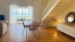 Ampla sala de estar com mesa de jantar, cadeiras, sofá, escada e vista para o exterior através de uma porta de vidro.