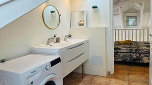 Quarto compacto com máquina de lavar, cama com colcha florida, mesa de cabeceira pequena e espelho.