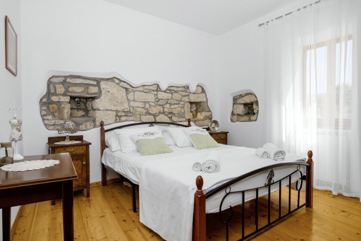A bedroom with a stone headboard, a wooden bed frame, a bedside table, and a window with sheer curtains.