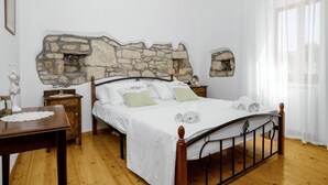 Un dormitorio con cabecera de piedra, estructura de cama de madera, una mesita de noche y una ventana con cortinas traslúcidas.