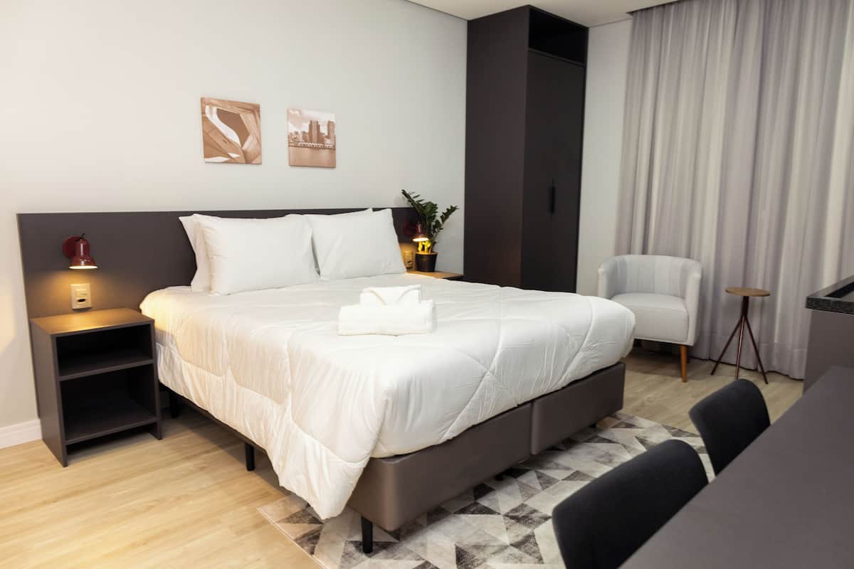 A modern hotel room with a large bed, a bedside table with a lamp, a chair, and a small desk area.