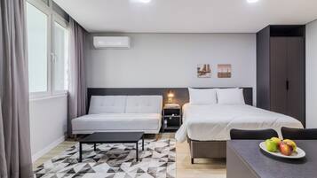A modern hotel room with a bed, a sofa, a small table, and a desk. There is a window with curtains, a wall-mounted air conditioner, and a nightstand with a lamp.