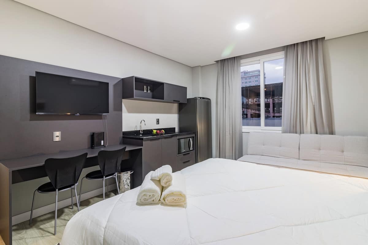 A hotel room with a bed, a desk with a chair, a TV, a microwave, and a window with curtains.