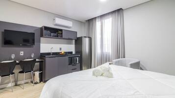 A modern hotel room with a bed, a desk, a kitchenette, and a TV.