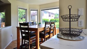 A dining area with a wooden table and chairs, a black metal basket on a marble countertop, and a view of a residential neighborhood through large windows. - Wonderful 4-bdrm, 2.5 bath home in Renton,15 min to Seattle,2 min to Coulon Park (Renton)