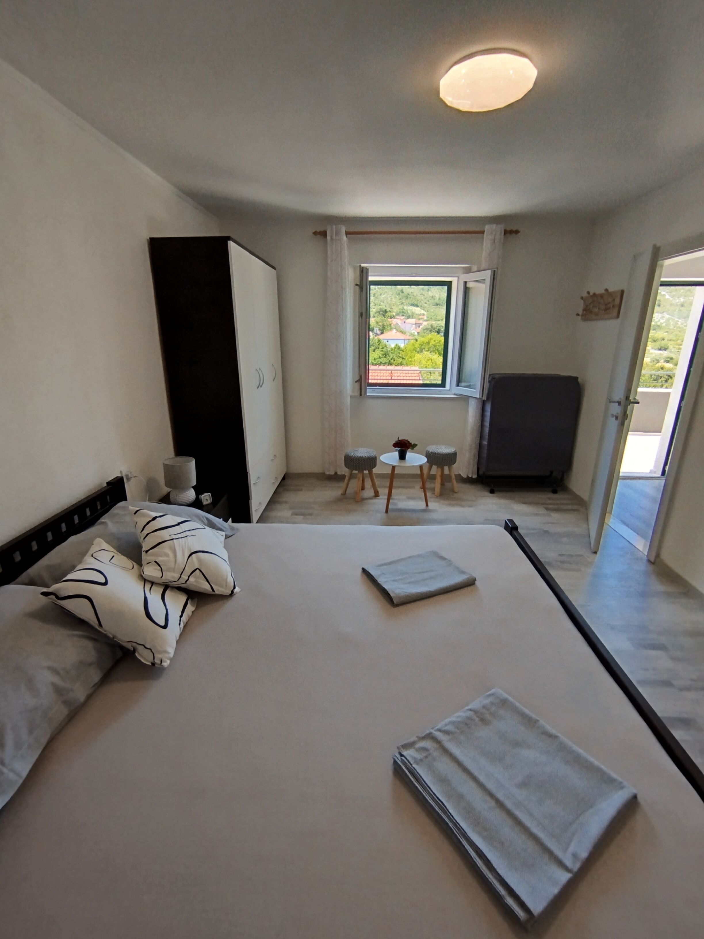 A bedroom with a bed, pillows, a small table with a vase, a chair, a wardrobe, and a window with a view of greenery.