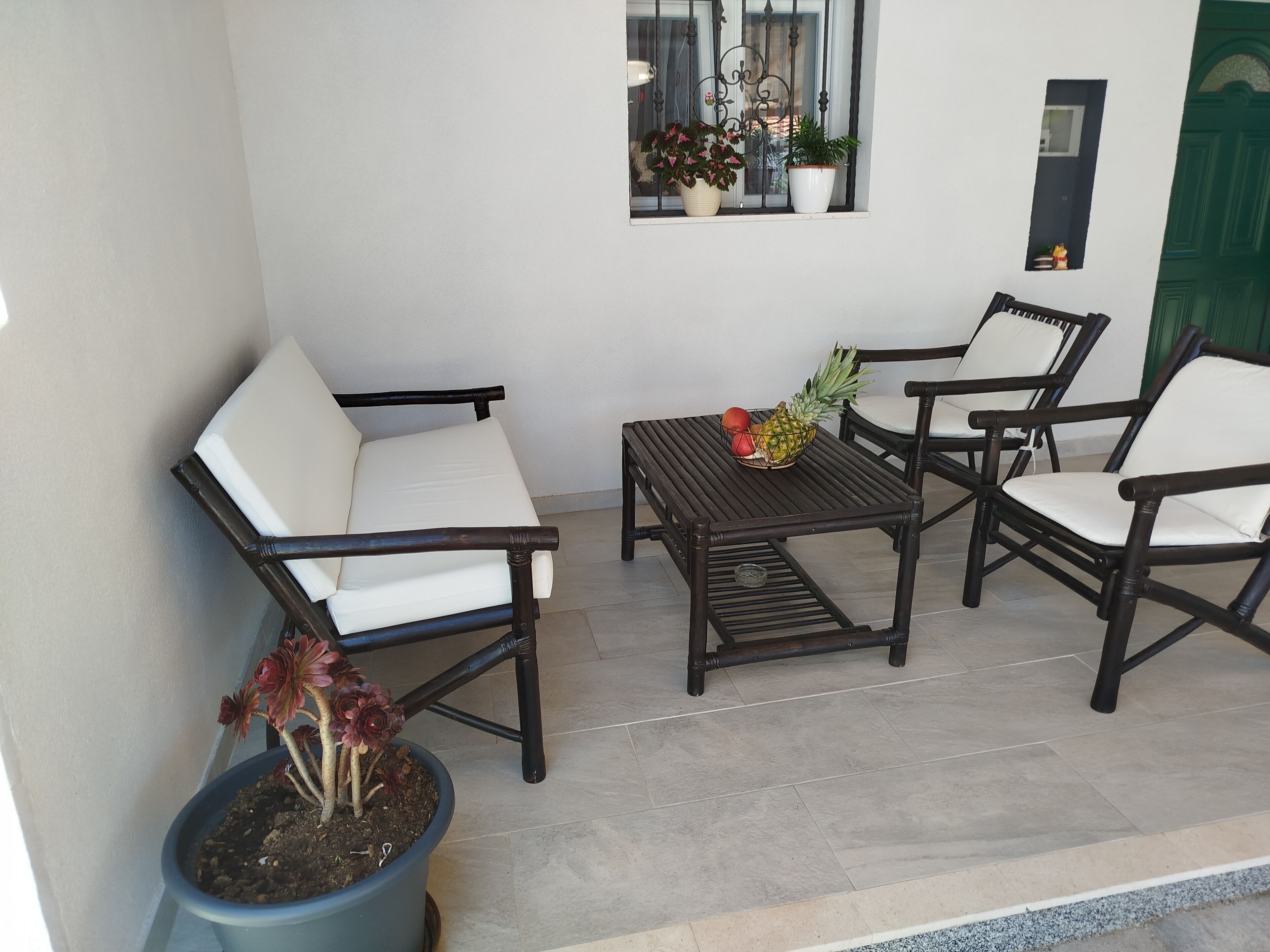 A patio area with two white cushioned chairs, a wooden table with fruit, and a potted plant.