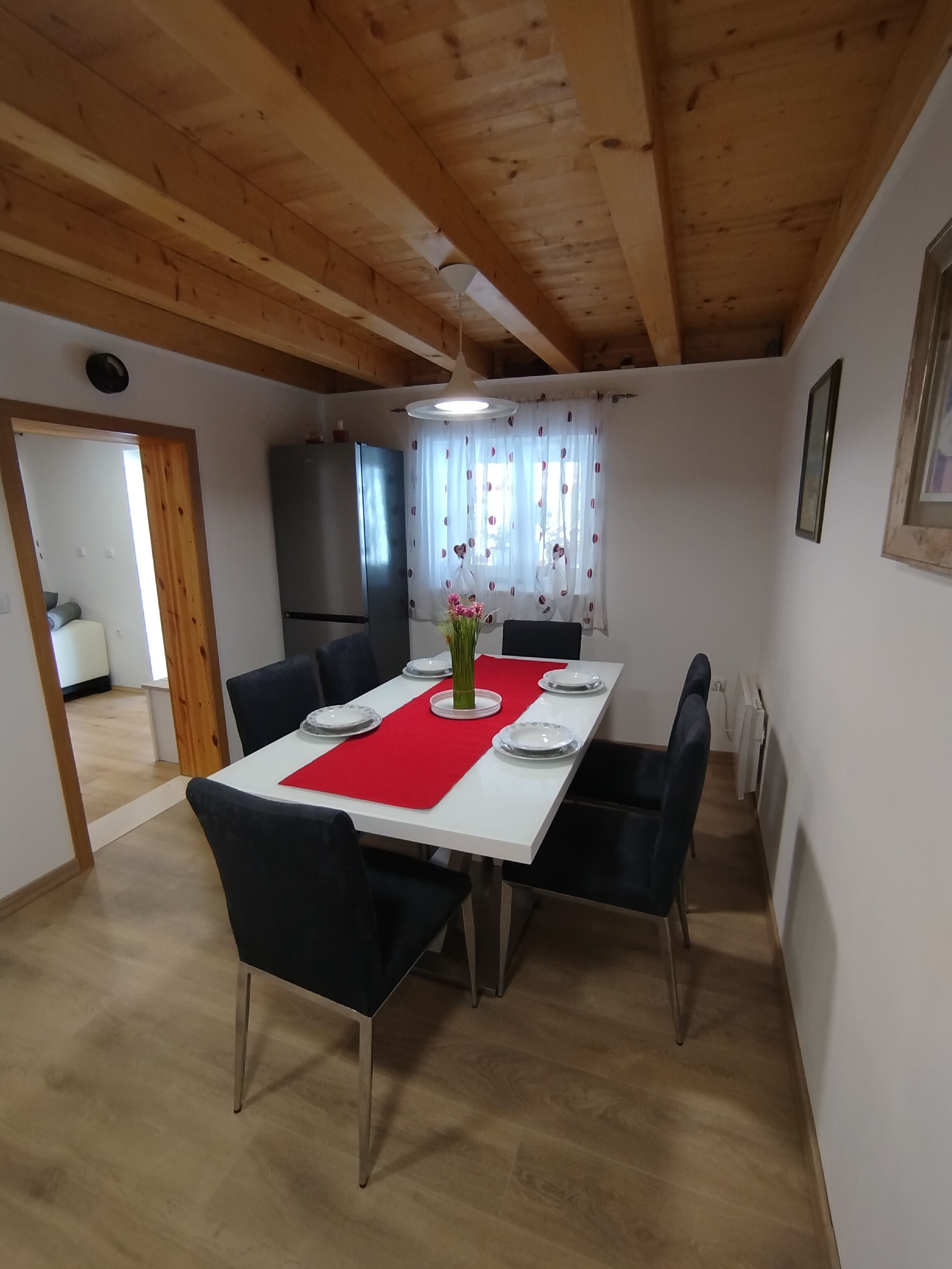 A dining area with a wooden ceiling, a white table with red placemats, black chairs, and a vase of flowers.