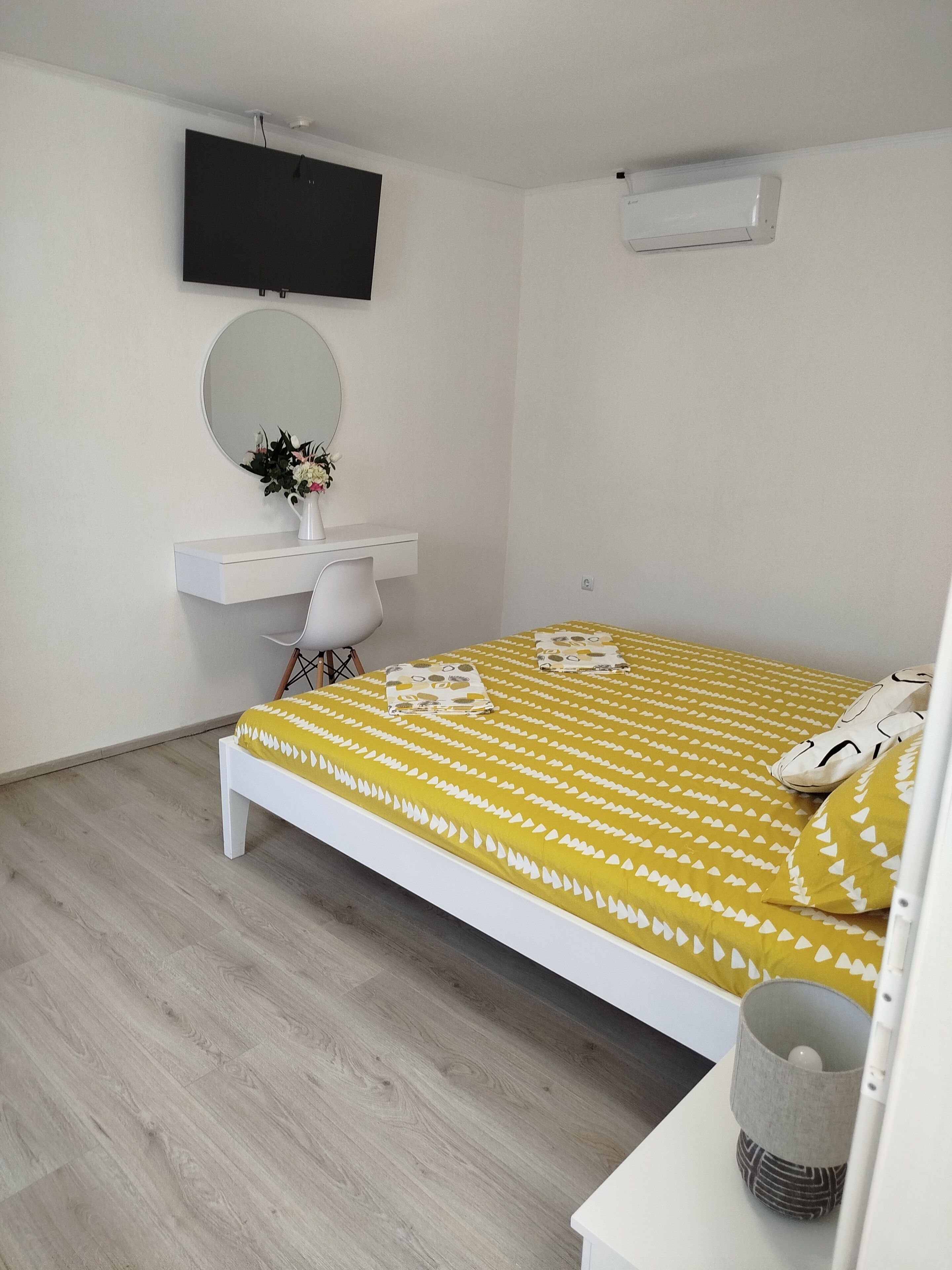 A bedroom with a bed, a desk with a chair, a round mirror, and a television on the wall.