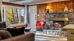 A cozy living room with a stone fireplace, wooden walls, and a view of trees through a large window. - 3BR Cozy Cottage, AC, BBQ, Fast Wi-Fi, Fireplace, Firepit, Walk to Beach (Pocono Lake)
