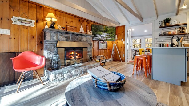 A cozy living room with a stone fireplace, wooden walls, a red chair, and a dining area with orange stools. - 3BR Cozy Cottage, AC, BBQ, Fast Wi-Fi, Fireplace, Firepit, Walk to Beach (Pocono Lake)