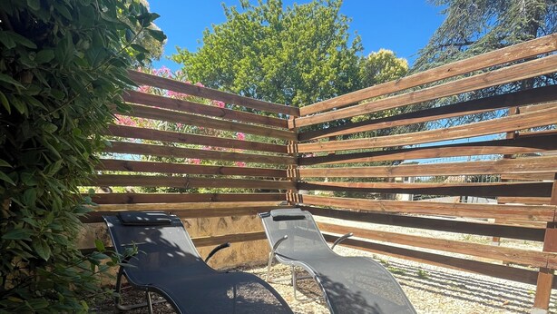 A wooden pergola with two black lounge chairs on a gravel surface.