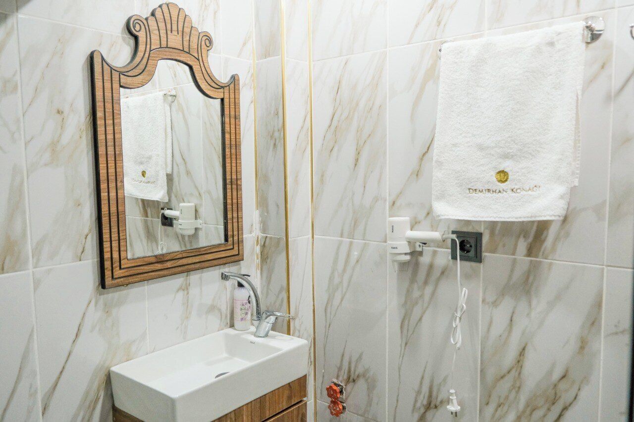Un baño con pared de mármol, espejo con marco de madera, lavamanos blanco y un perchero con toalla blanca.
